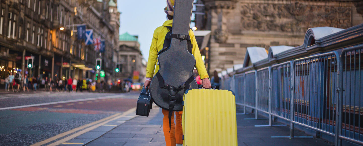 A musician with a guitar case on their back walks down a street in Edinburgh while pulling a suitcase
