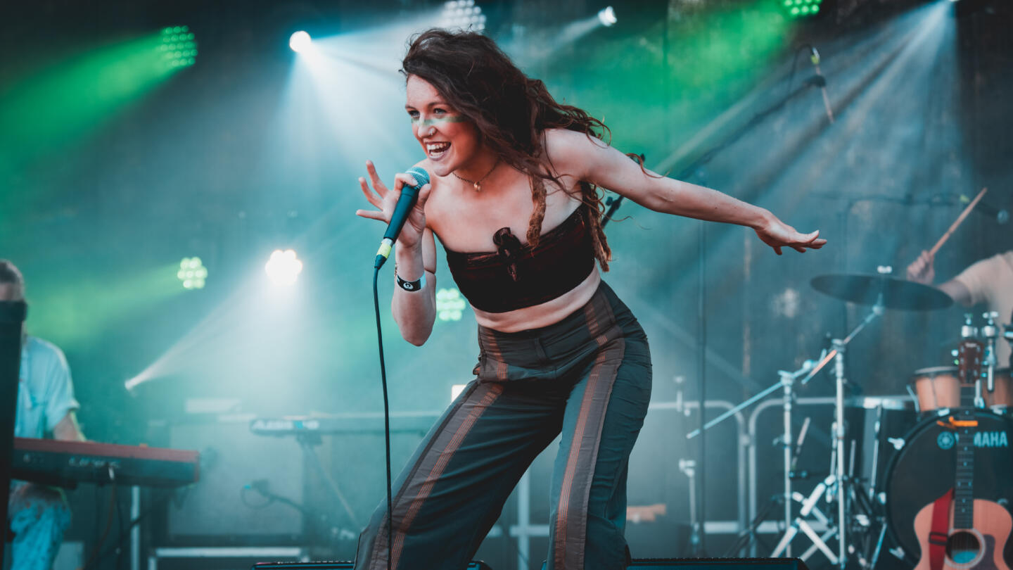 A woman singing into a microphone on a festival stage, surrounded by her band