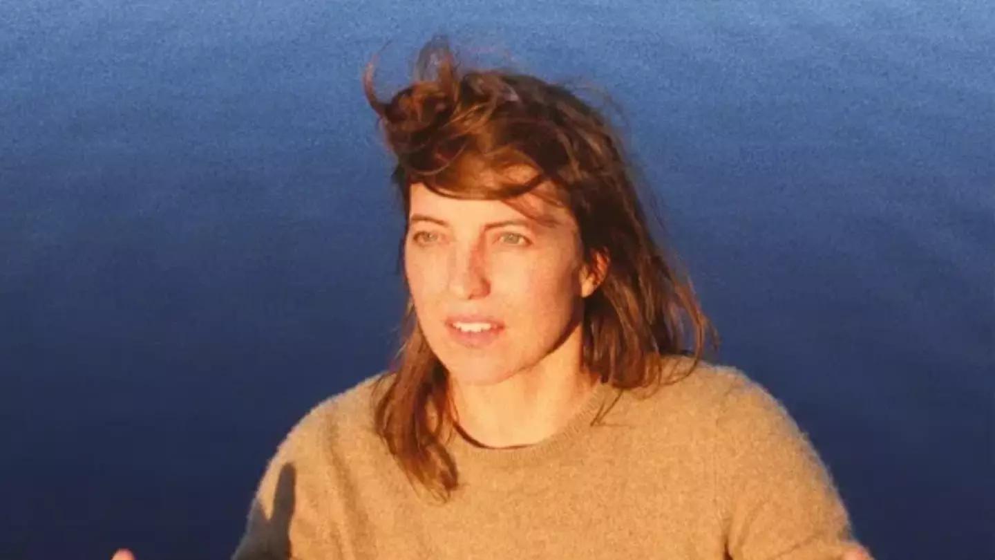 Rozi Plain standing in front of a body of water. She has thrown a baseball cap in the air.