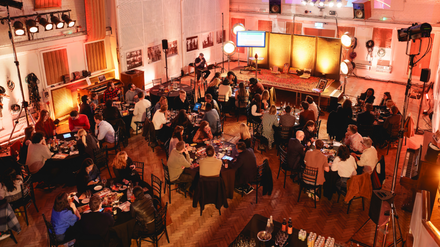 A large event hall with multiple round tables filled with attendees, illuminated by event lighting, and a podium set on a stage at the front of the room.