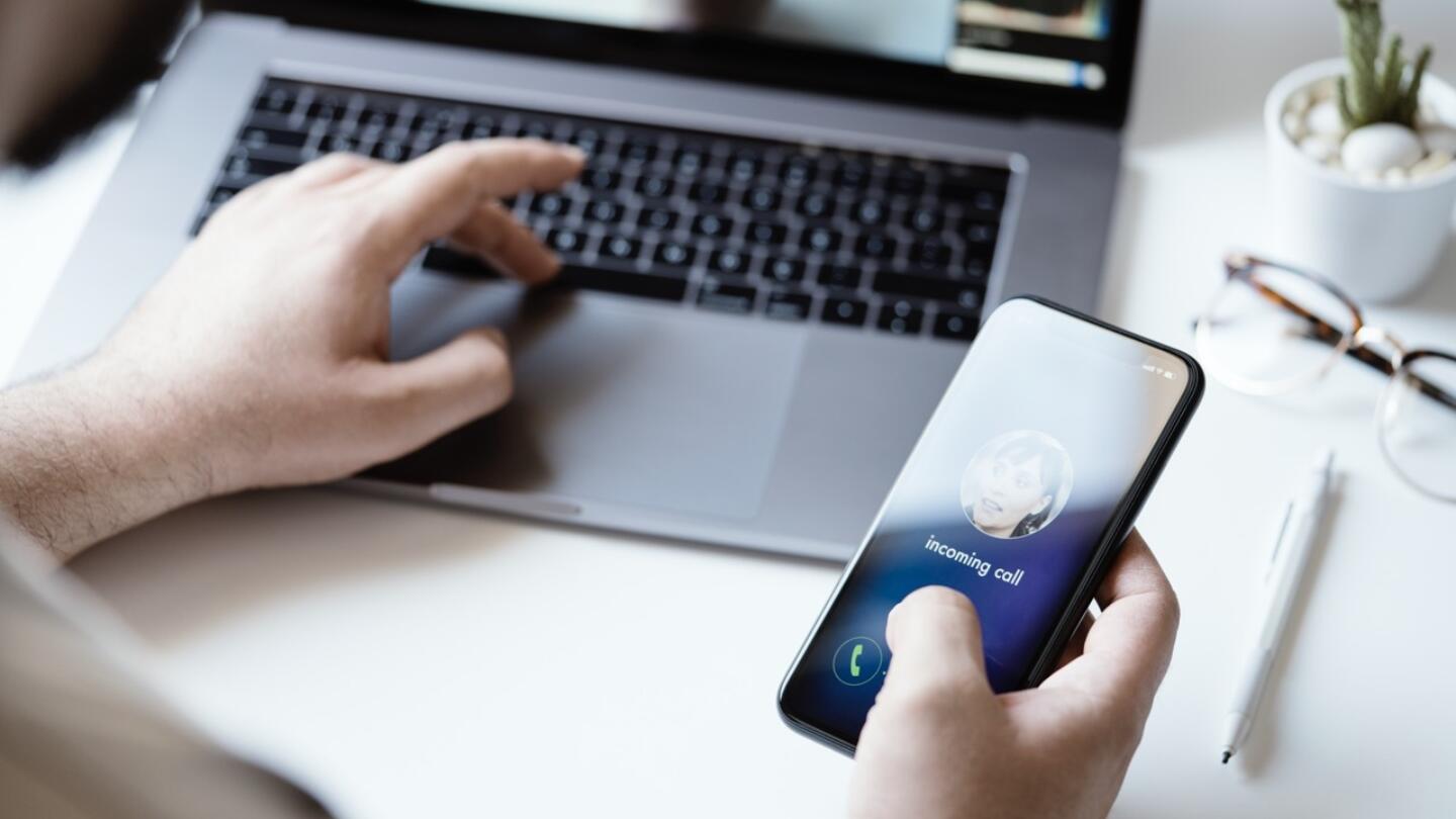 A person holds their phone in one hand and types at a laptop with the other