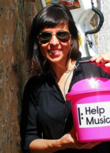 Jess Cox outside leaning against a wall holding a collection bucket for Help Musicians