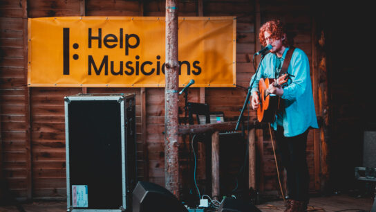 A guitarist performs in front of a large Help Musicians banner
