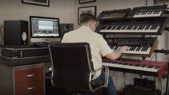 Tom from Rhumba Club sat facing away from the camera playing on a stack of keyboards on his desk
