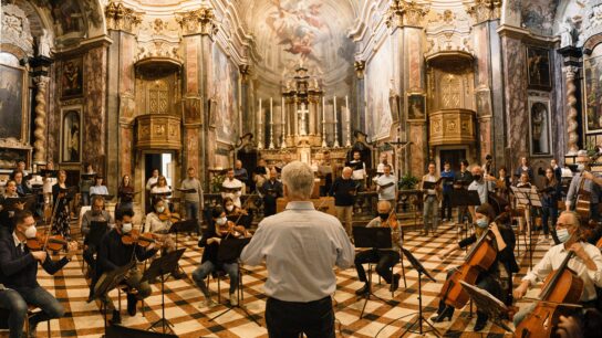 A full orchestra sit in an old chapel being led by a conductor