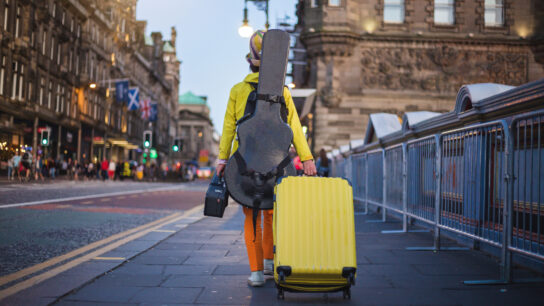 A musician with a guitar case on their back walks down a street in Edinburgh while pulling a suitcase