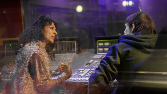 A woman and a man are sat together at a sound desk. The woman appears to be offering advice to him.