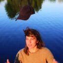 Rozi Plain standing in front of a body of water. She has thrown a baseball hat in the air. logo